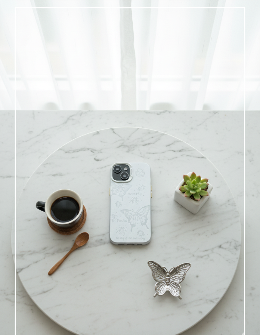 Phone, coffee cup, butterfly decor, and small plant on a marble surface with white curtains in the background.